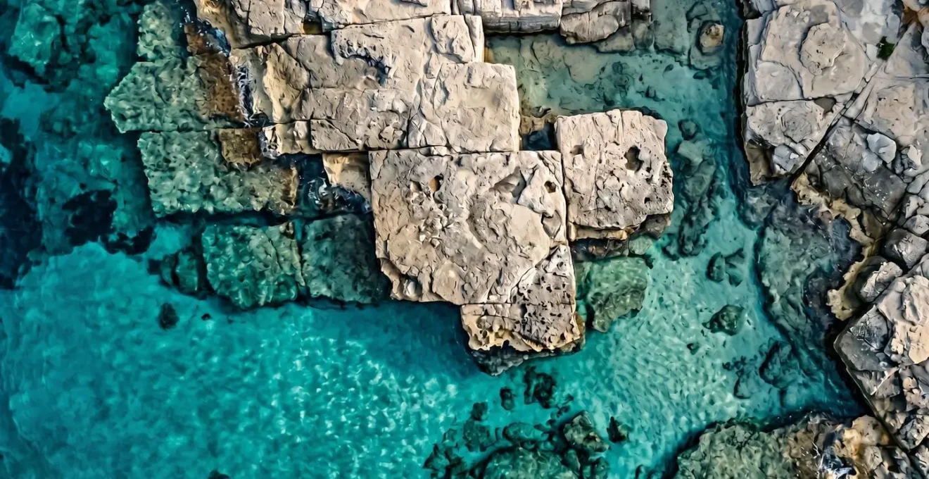 Vista aerea zenitale di rocce piatte grigio-beige circondate da acqua turchese cristallina con un piccolo asciugamano colorato come unico elemento umano